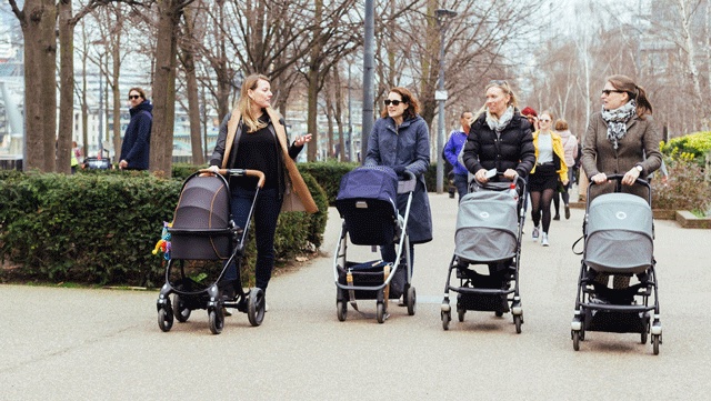 a group of first time moms pushing strollers in the park in a city with coats on