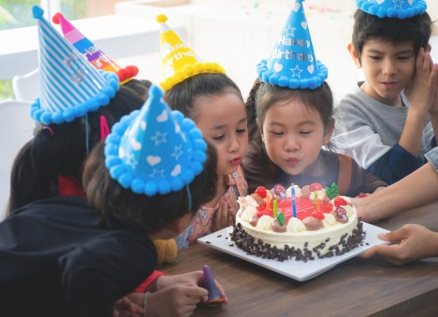 Little kids crowd around a birthday cake at home while one blows out a candle