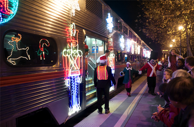 People walking by a train from Caltrain decorated with Christmas lights and festive displays like Nutcrackers and reindeer on the outside of the train.