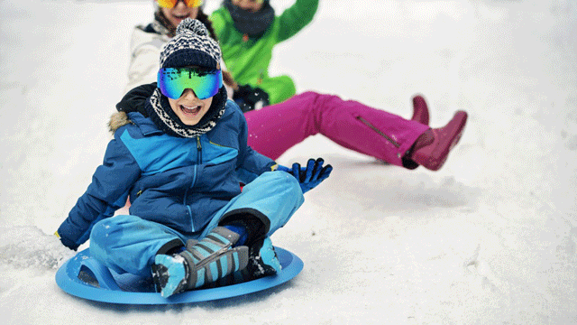 A child in blue winter gear sleds down a snowy hill near Portland followed by his two friends