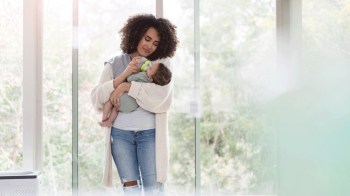 a mom stands in front of a window feeding her baby with a bottle