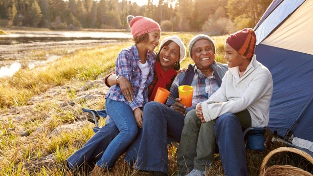 a family in hats hold mugs around a campfire with the sun setting in th background at a campgrounds near portland, oregon