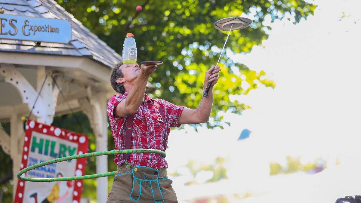 a man spins a plate, hula-hoops and balances a bottle of water on his nose at a boston festival