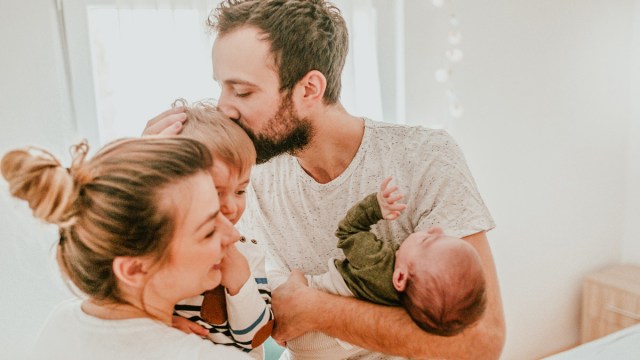 A mom holding a toddler while a dad, holding their new baby, kisses his head
