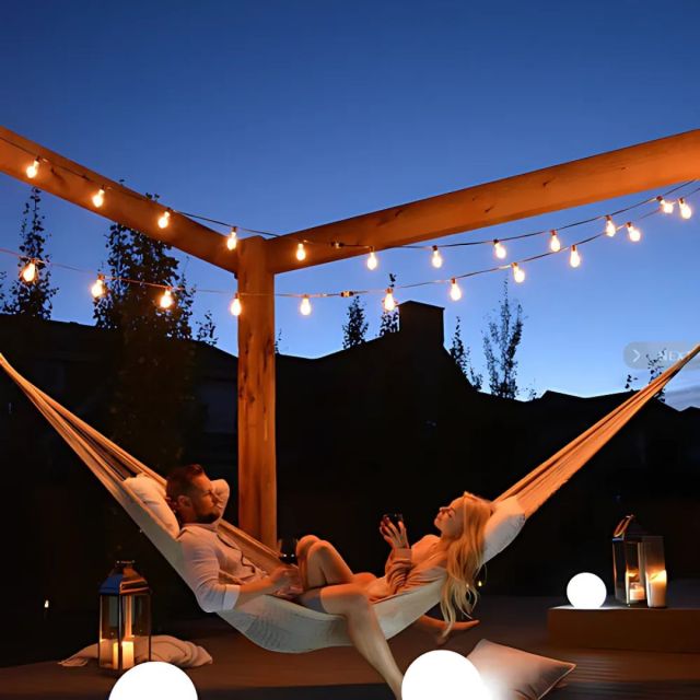 a couple lounging in a backyard hammock attached to a pergola wrapped in outdoor lights